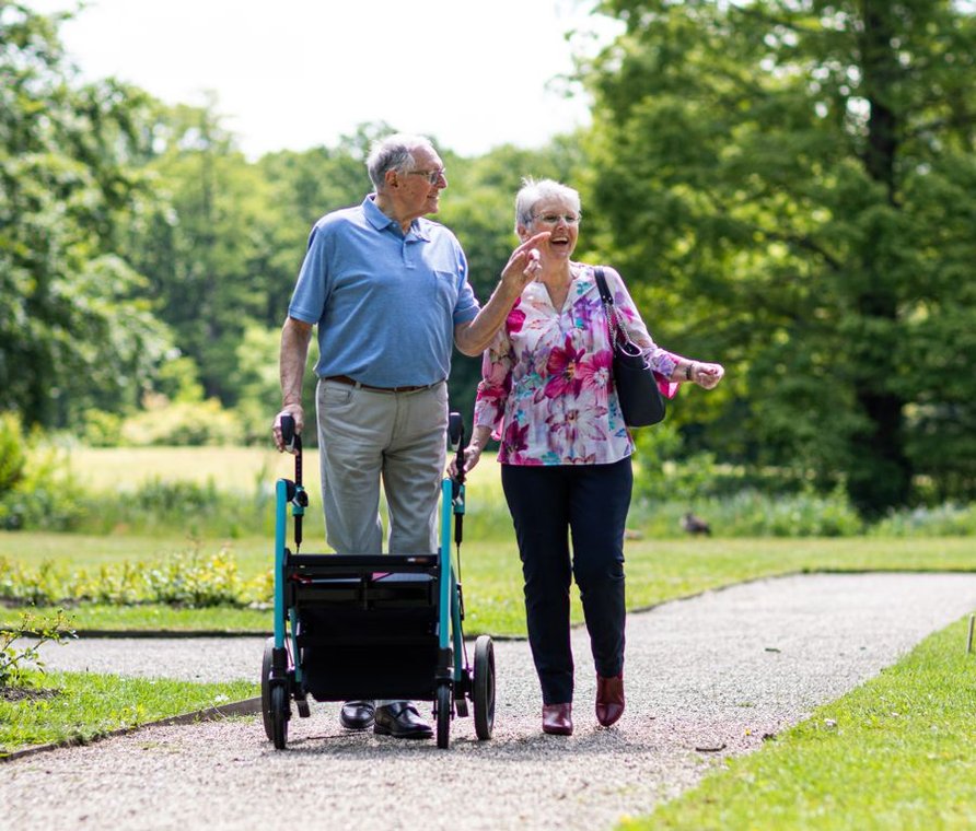 Older couple walking outdoors on a garden path, one using a rolling walker