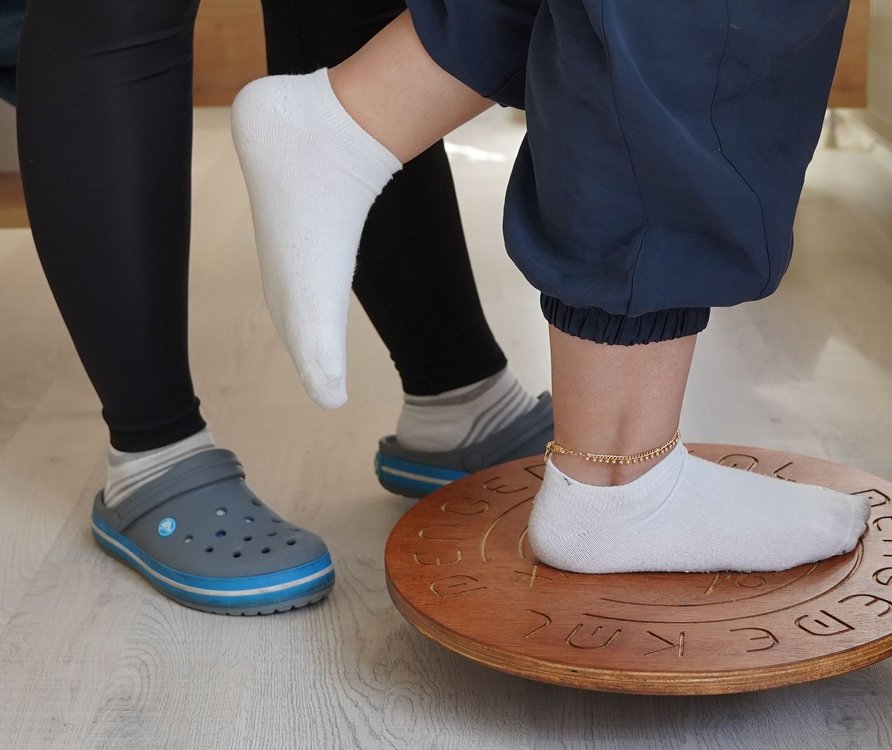 Patient balancing on one foot on a wooden wobble board with a therapist standing close beside them